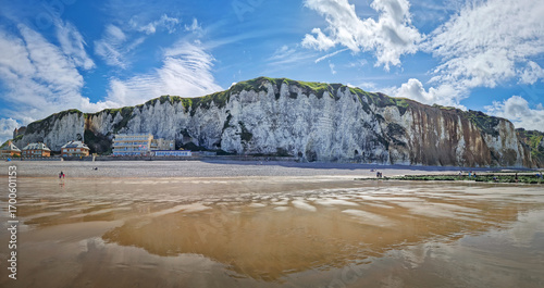 Panoramic view of Dieppe beach in Normandy, France. Majestic white chalk cliffs rise above a pebble shore, with the low tide revealing a vast expanse of wet sand reflecting the blue sky