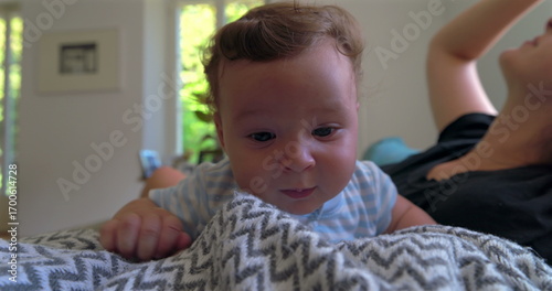 Baby lying on a blanket, looking slightly frustrated and emotional, in a warm home setting with natural light, while mother observes in the background
