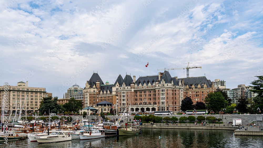 Naklejka premium Victoria inner harbour causeway glows warmly at sunset with shimmering water reflecting the colourful sky, boats gently bobbing, and the city's historic skyline creating a serene, picturesque scene.