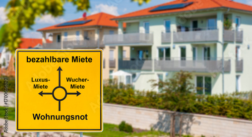 Vor einem modernen Mehrfamilienhaus steht ein symbolisches Schild mit der Aufschrift von der Wohnungsnot zur bezahlbaren Miete - FOTOMONTAGE