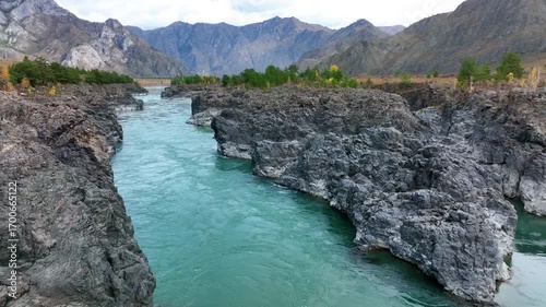 An aerial view of a vibrant turquoise river flowing through a narrow, rocky gorge with a rugged shore, surrounded by mountains with autumn foliage
