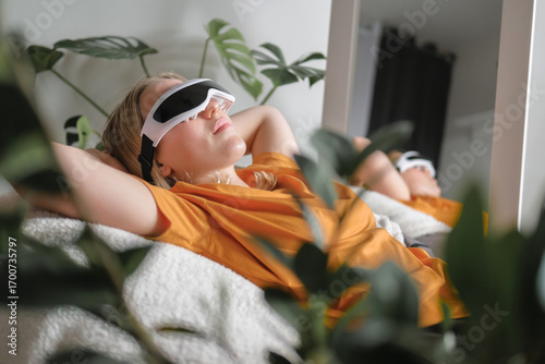Gen Z woman unwinds  home smart eye massager?wellness tech  recovery, mindfulness and stress relief. Cozy greenery and soft light set a self-care, digital-detox mood, promoting balance, calm wellbeing