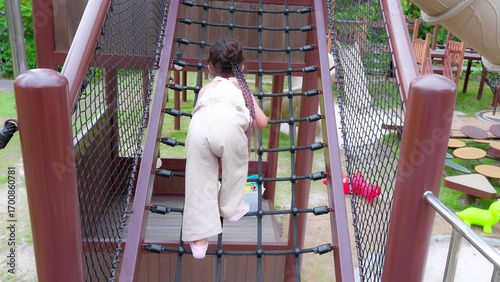 Happy Young Asian child girl climbing a rope bridge at a playground, showcasing outdoor play and adventure.