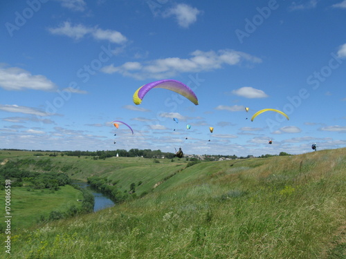 Colorful paragliders soaring above green hills and a river on summer day  