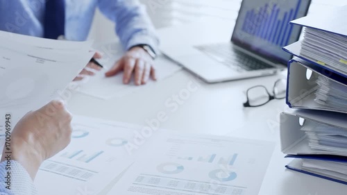 Office workers or auditors are analyzing financial reports and checking documents while sitting at white table with a stack of blue folders in the foreground. Audit and taxes theme in business