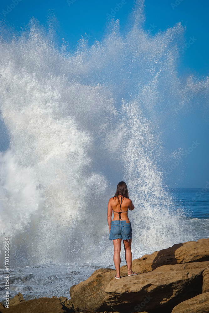 Fototapeta premium PORTRAIT: Young woman is admiring a huge wave crashing against rugged rocky Moroccan shore sending up a large white splash of foam and spray. Beach walk with dramatic scenery of mighty Atlantic Ocean