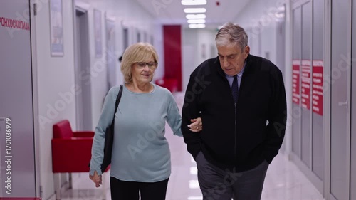 An older couple walking together in a brightly lit hospital hallway, with concern
