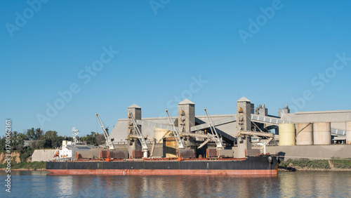 Cargo ship loading grain at an industrial port facility on a sunny day. Showcasing global trade & logistics
