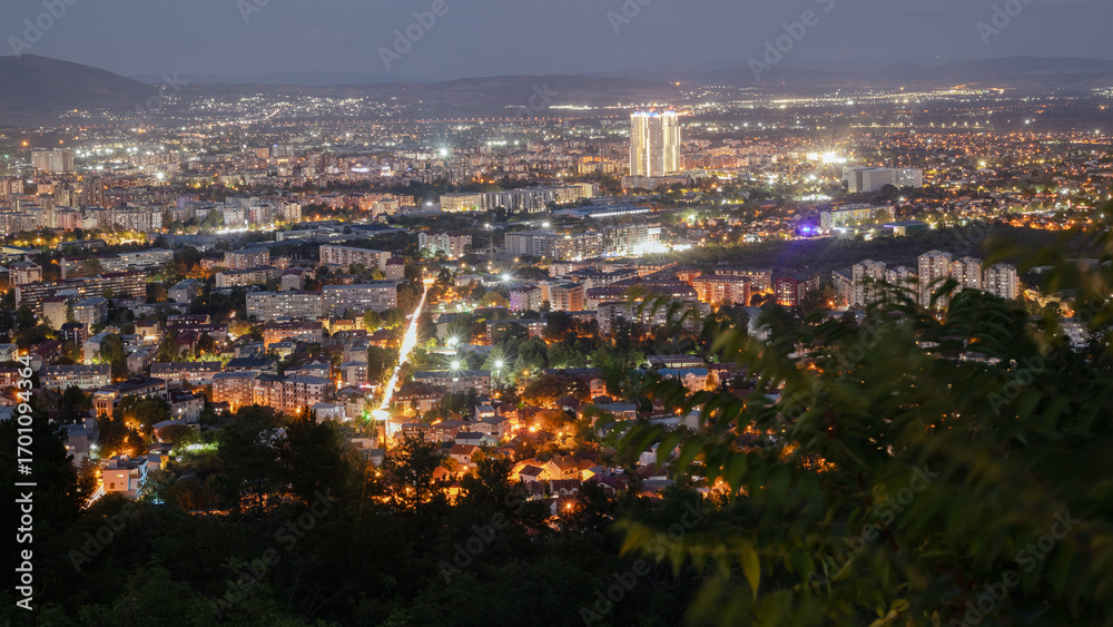 Naklejka premium an aerial view of the city of skopje at night, showing the urban landscape with illuminated buildings and streets under a twilight sky. the city lights create a vibrant and warm glow