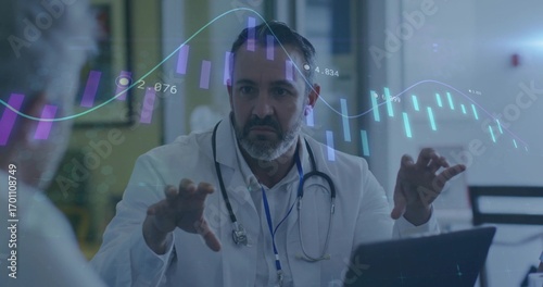 Gesturing male doctor explaining laptop data in hospital room, with stethoscope and floating charts