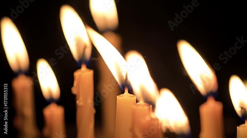 Multiple lit candles with bright, flickering flames casting a warm glow in a dark, solemn setting, creating a peaceful atmosphere.