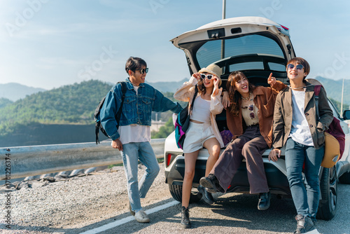 Happy Young Asian man and woman friends road trip travel countryside on summer holiday vacation. Generation z people enjoy outdoor active lifestyle sitting on EV car trunk and looking beautiful nature
