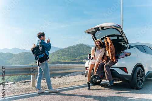 Happy Young Asian man and woman road trip travel by EV car countryside on summer holiday vacation. Generation z people friends sitting on car trunk and using mobile phone taking picture together.