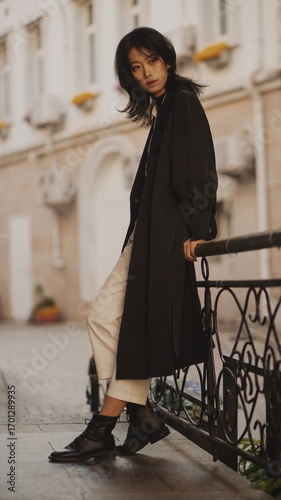 Stylish woman in a black coat and white pants poses against a vintage building backdrop.