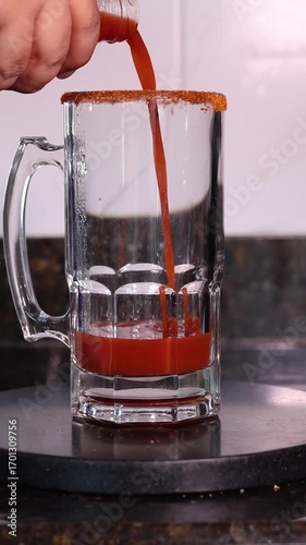 Tomato and clam juice being poured into a large beer mug with a spicy rim, ready for a michelada cocktail