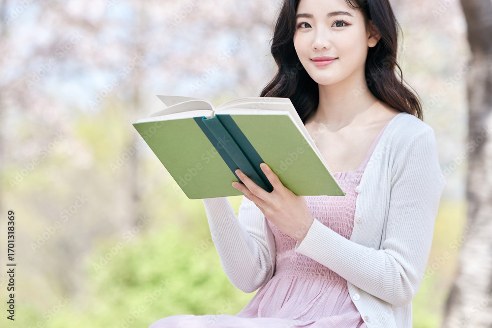Obraz premium Young Asian female reading book outdoors in spring park, wearing pink dress and white cardigan