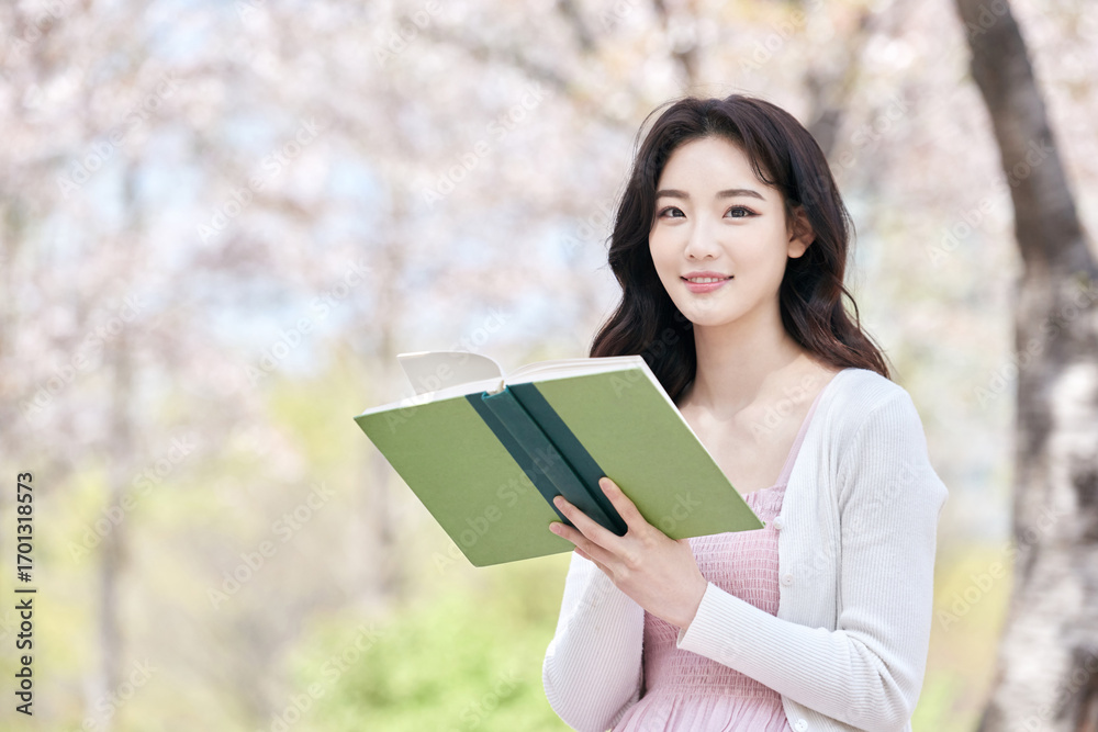 Obraz premium Young Asian female reading book outdoors in spring park, wearing pink dress and white cardigan