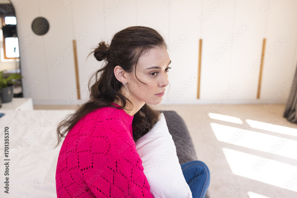 Naklejka premium Woman sitting on bed holding white pillow in bright modern bedroom, wearing pink knit sweater