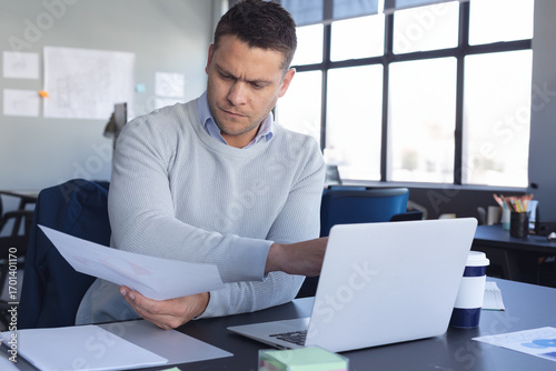Bilde på lerret Businessman reviewing documents at desk with laptop and coffee cup in office