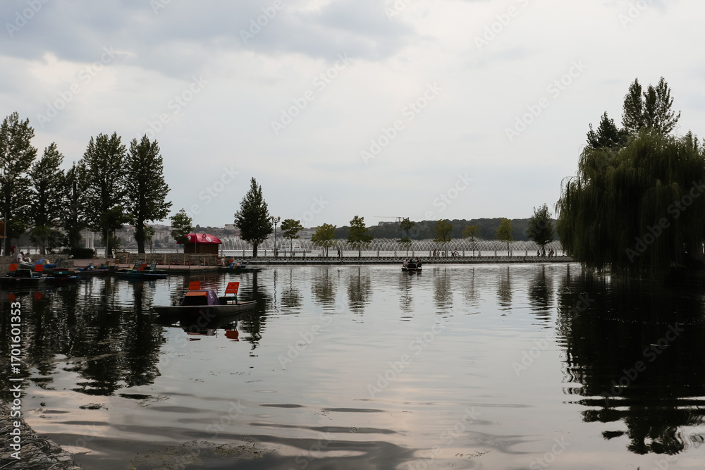 Fototapeta premium A serene lake with paddle boats and people enjoying a calm day, with the lush trees and a scenic pier reflecting on the tranquil water's surface.
