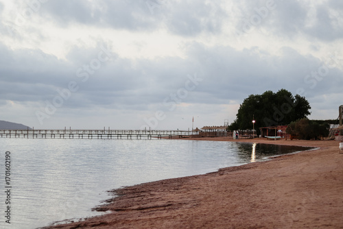 Wallpaper Mural A calm beach at dusk with a gentle curve of sand leading to the water, a distant pier, and a few buildings under a dramatic, cloudy sky. The soft light reflects on the still water Torontodigital.ca