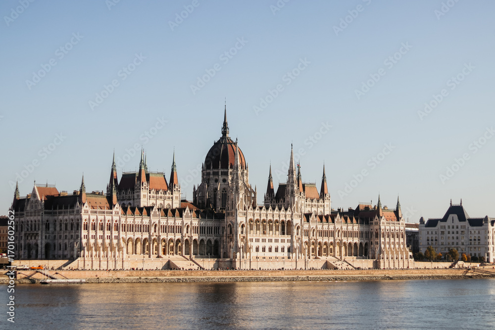 Obraz premium The stunning Hungarian Parliament Building in Budapest, Hungary, stands proudly on the bank of the Danube River, showcasing neo-Gothic architecture against a clear blue sky.