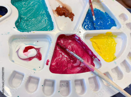 A white watercolor palette mixing plastic tray with several compartments , containing various acrylic for mixed colors such as red, yellow, blue, and green. Focus on red with a paintbrush.