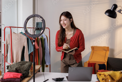 Young Asia lady seller look at phone camera live stream use laptop answer follower comment introduce sweater new collection new arrival on her chanel at studio. Woman fashion online sme shop concept.