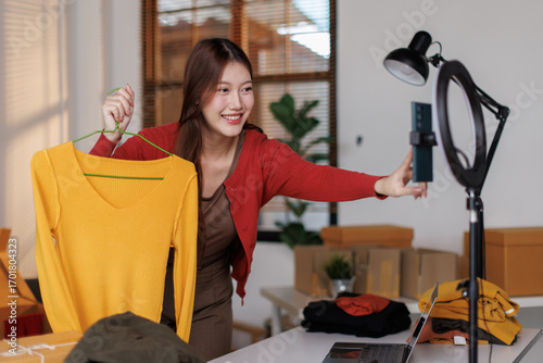 Young Asia lady seller look at phone camera live stream use laptop answer follower comment introduce sweater new collection new arrival on her chanel at studio. Woman fashion online sme shop concept.