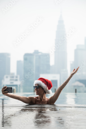 Photography Woman wearing santa hat taking selfie in rooftop pool overlooking kuala lumpur s