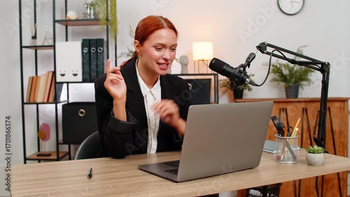 Young businesswoman at home office talks into studio microphone recording podcast blog video laughing. Freelancer girl by table near laptop shares ideas engaging audience and enjoying fun joyful work.