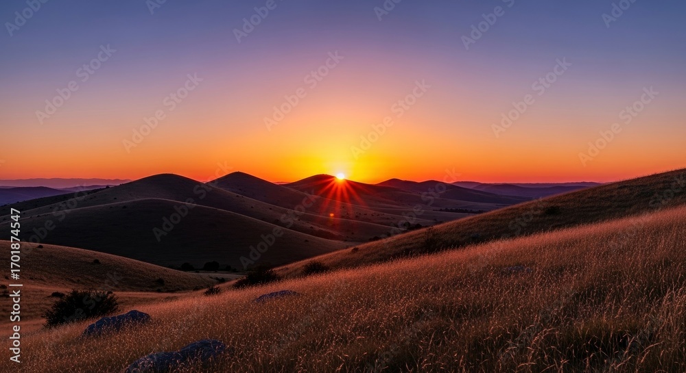 Obraz premium Sunset between rolling hills with radiant sunbeams over a grassy landscape