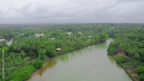 Aerial drone shot flying over backwater canal to buildings in the middle of coconut trees with domed roofs of the Taj hotel under monsoon clouds showing Kappil Bekal Kerala