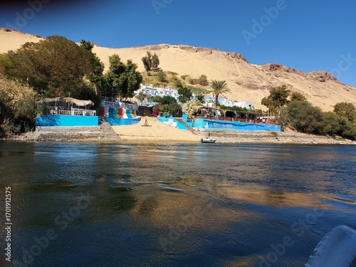 Colorful Nubian House on the Nile River, A vibrant view of a brightly painted house on the sandy banks of the Nile River. The building, a typical example of Nubian architecture.