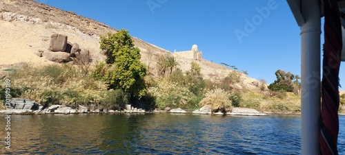 Scenic Riverbank on the Nile River, Aswan, A peaceful view from a boat on the Nile River, showing a lush, green riverbank contrasting with a barren, rocky hillside in the background.
