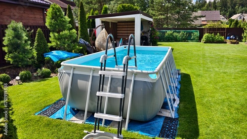 On the mowed grassy lawn stands a gray rectangular PVC frame pool with a ladder. The water in it is filtered. Nearby is a wooden gazebo, swings, bushes and trees. Sunny summer weather and blue sky