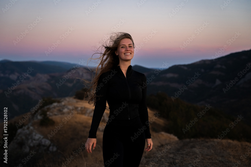 Naklejka premium Happy woman with tousled hair standing in mountains at sunset, female traveler smiling at camera and enjoying freedom outdoor, relaxing in nature in summer, active lifestyle