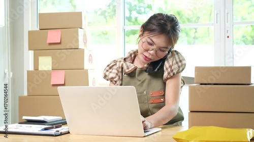 Young Asian female entrepreneur manages online business orders from home using smartphone, laptop and e-commerce work package.