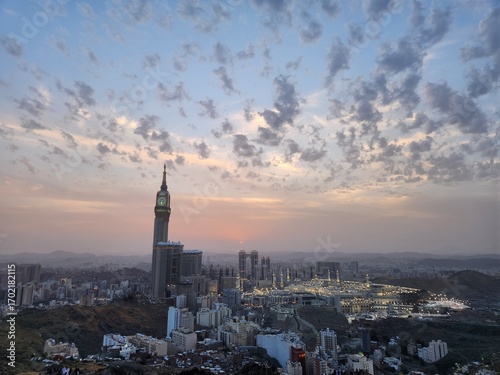 View of Mecca, Saudi Arabia, with the Abraj Al Bait Clock Tower prominently on the left. The cityscape includes numerous modern buildings and the surrounding rocky terrain of Jabal Khandamah. The sky 