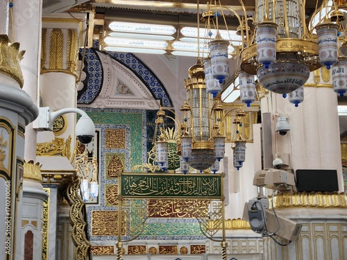 Ornate interior of Al-Masjid an-Nabawi in Medina, Saudi Arabia, known as the Prophet's Mosque. The image shows intricately designed chandeliers with calligraphic inscriptions and golden accents hangin