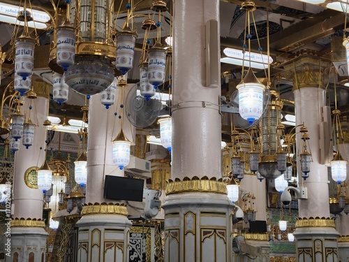 Ornate interior of Al-Masjid an-Nabawi in Medina, Saudi Arabia, known as the Prophet's Mosque. The image shows intricately designed chandeliers with calligraphic inscriptions and golden accents hangin