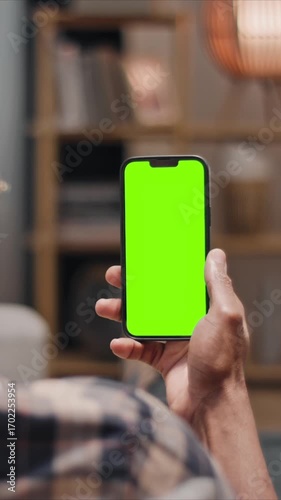 Man holding smartphone with green screen close up, mobile phone in hand, using chroma key mockup indoors at home. Male person browsing blank screen device in vertical position.