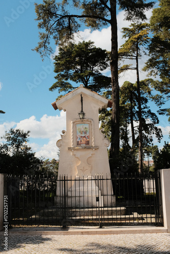 Catholic roadside shrine in Fatima Portugal with tiled Virgin Mary image and metal fence surrounded by pine trees under clear blue sky religious monument and local devotion site. High quality photo