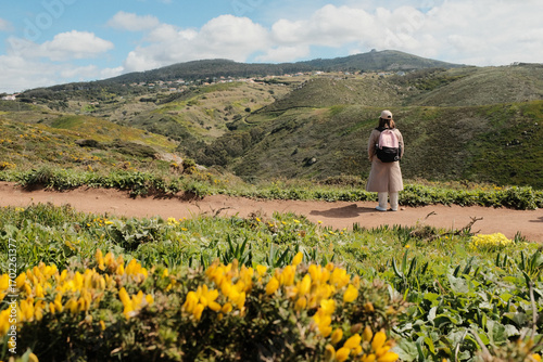 Woman enjoying springtime flower field on scenic hiking trail with mountain hills in background at Cabo da Roca Sintra Portugal nature walk with wild yellow blooms and blue sky. High quality photo