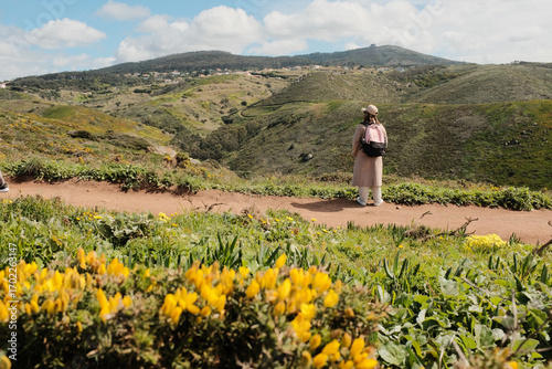 Woman enjoying springtime flower field on scenic hiking trail with mountain hills in background at Cabo da Roca Sintra Portugal nature walk with wild yellow blooms and blue sky. High quality photo