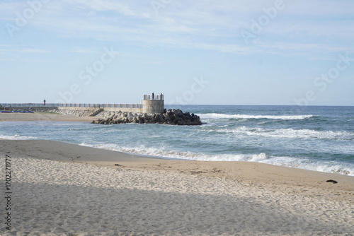 Concrete breakwater with small observation deck on sandy beach and ocean waves, coastal seascape landscape with sea horizon under clear blue sky