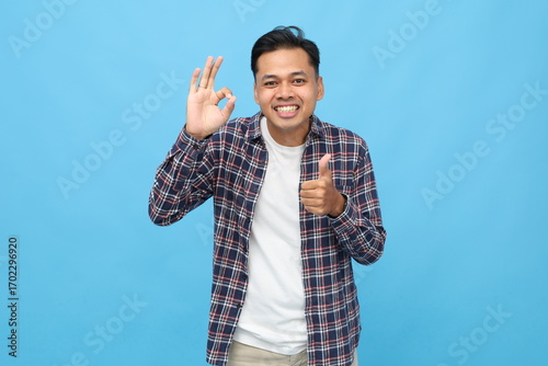 Handsome young Asian man wearing casual clothes, showing ok sign hand gesture and thumbs up, smiling looking at camera, feeling satisfied, standing isolated on blue background.