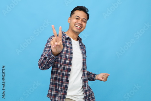 Handsome young asian man in casual clothes, smiling and showing victory sign, raising two fingers v gesture and clenching fist, cheerful, happy looking at camera, standing isolated on blue background.