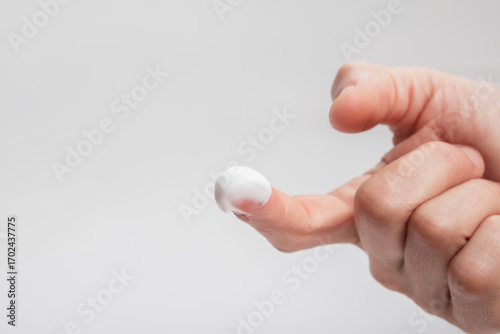 Hand with cream on finger against gray background. Close up of woman finger with a drop of white lotion or rub. Skin care moisturizer or medical prescription application. Copy space. Selective focus.