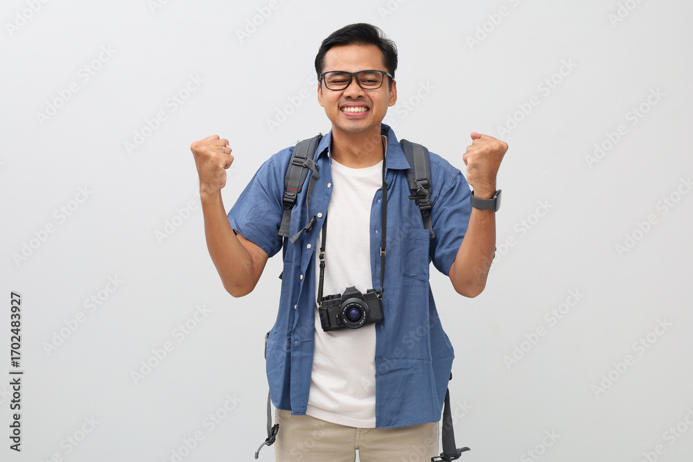 Fototapeta premium A young asian man wearing casual clothes, backpack, camera hanging on neck, making a gesture of celebrating victory by clenching his fist, smiling while closing his eyes, travel or vacation concept.